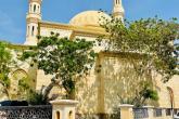 The Jumeirah Mosque sits in the sun under a blue sky with a couple trees in front. There are two minarets that rise up over a domed roof.