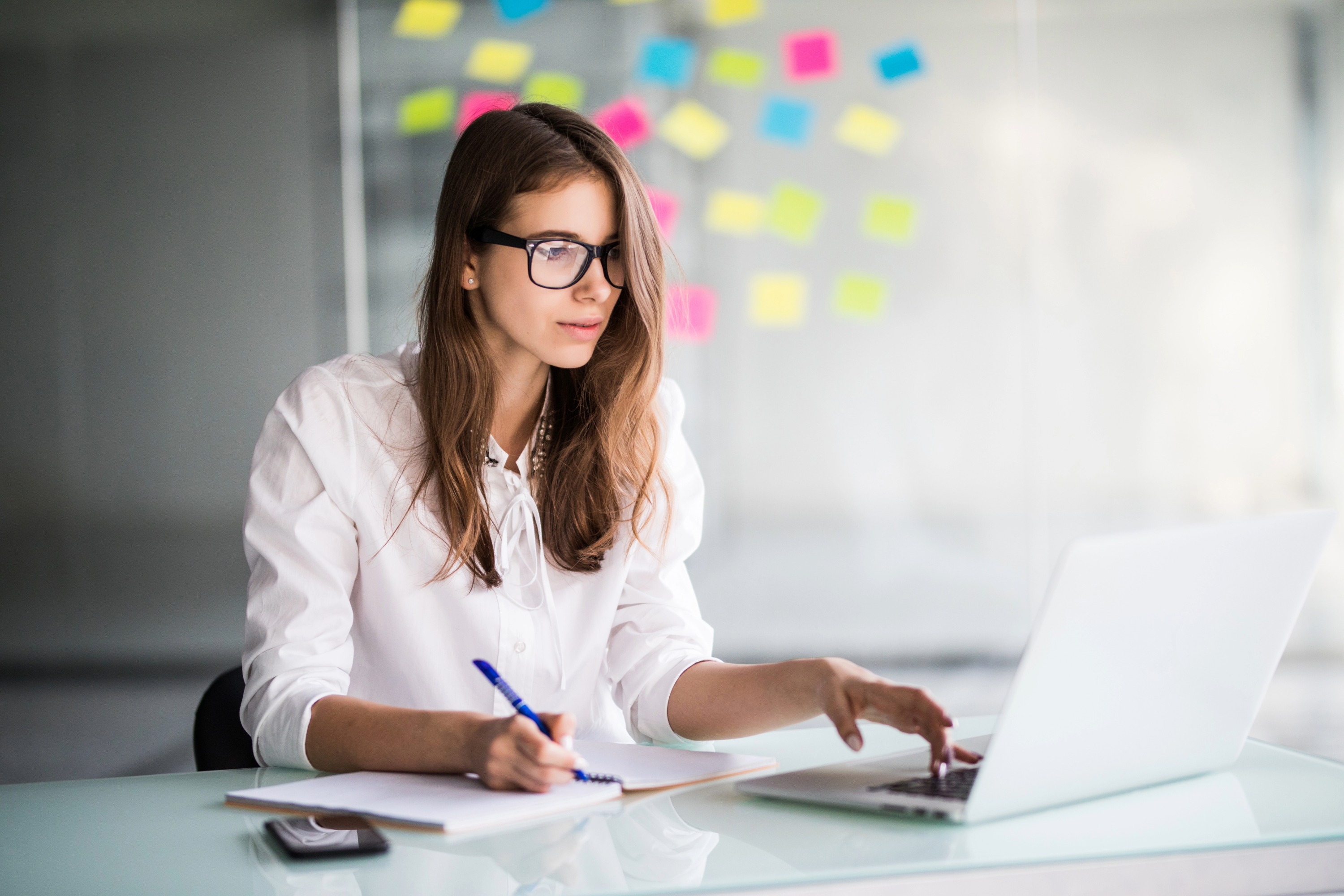 Smiling young business woman sitting and writing something by the table looking on laptop
