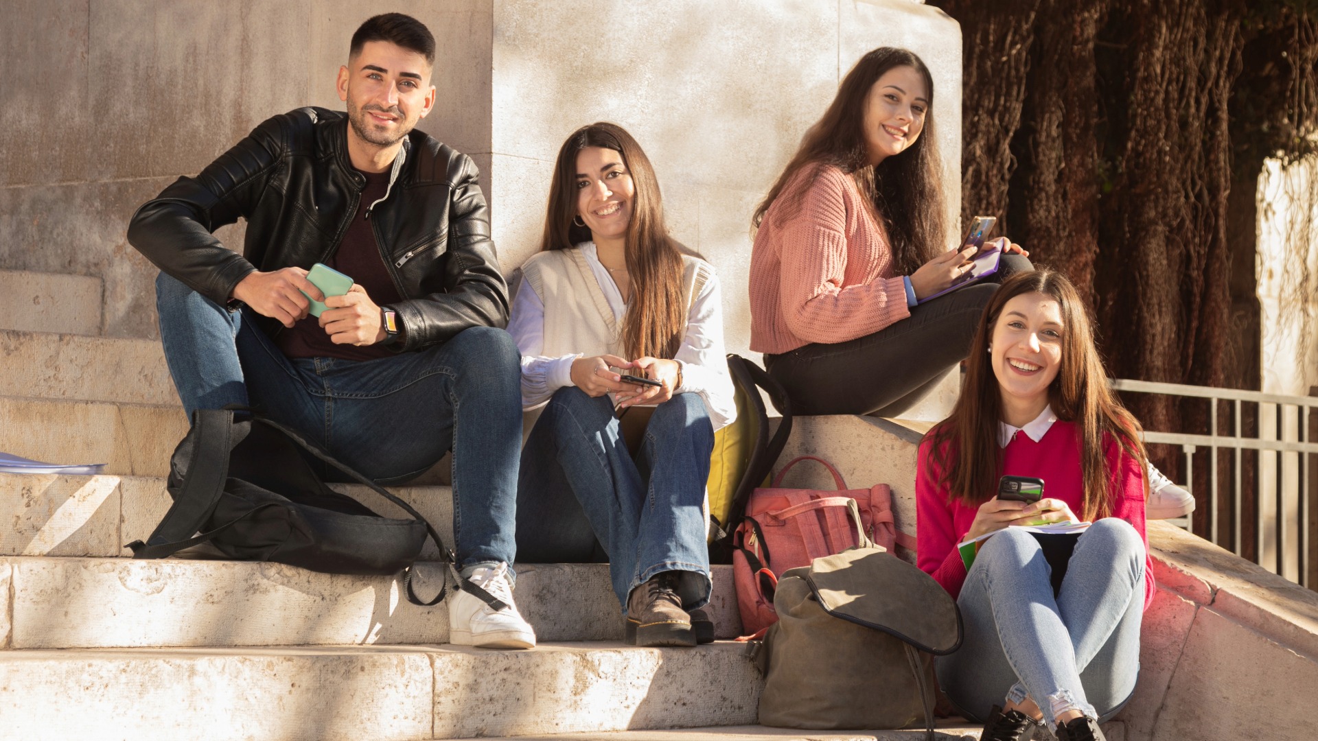 Four students looking at the camera and smiling while holding their smartphones sitting on the stairs of the university