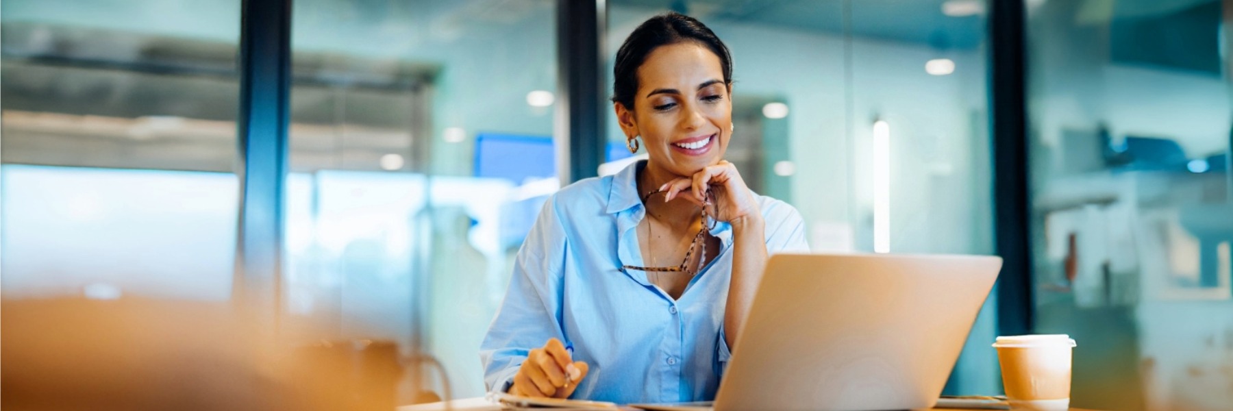 Smiling woman in office using laptop while holding glasses and working thoughtfully TOEIC