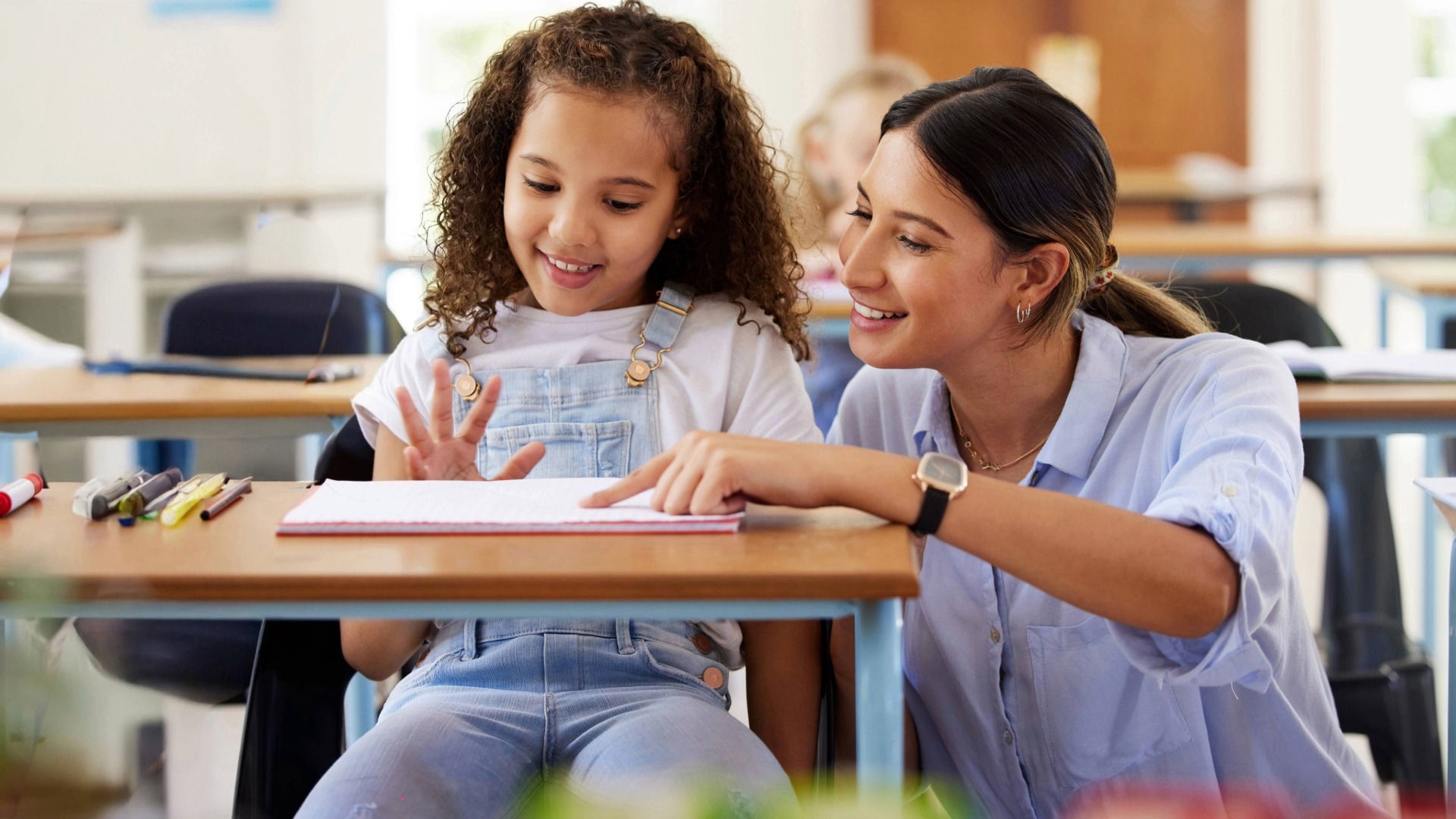 Teacher pointing to a book while a young student looks on attentively in a classroom setting, engaged in learning.