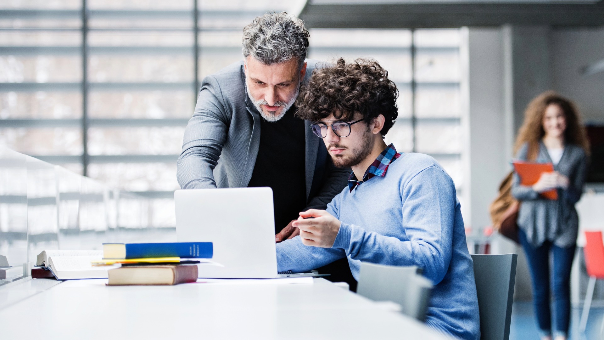 University professor helping student with final project, showing him research paper, book for thesis in library.