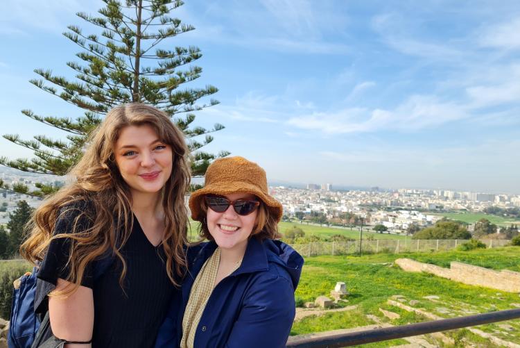 Two women stand a smile in front of a vista of green fields and city in the background.