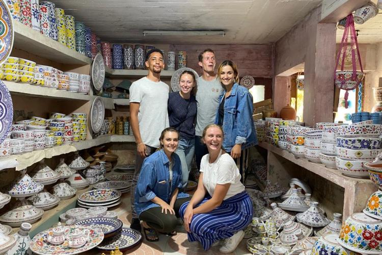 6 people pose for the camera in a pottery shop with finished products all around them on shelves waiting to be sold.