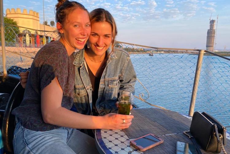 Two women sit together alongside the river drinking tea. The setting sun lights their smiling faces.