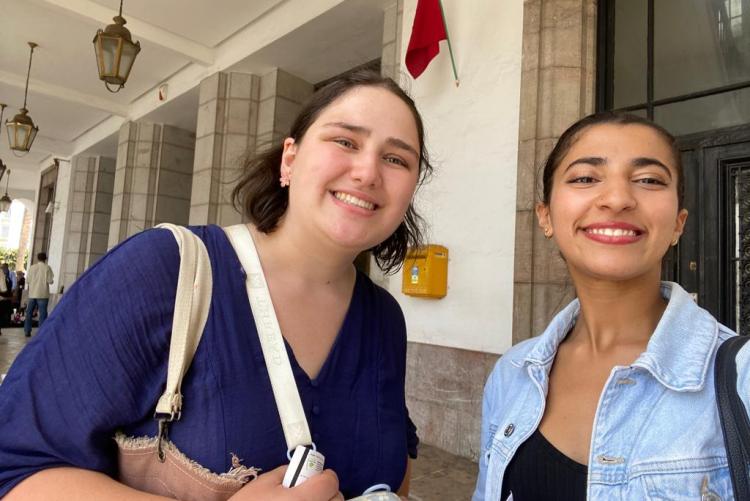 Two women pose together and smile for a selfie. They are both smiling and are standing in front of a large building.