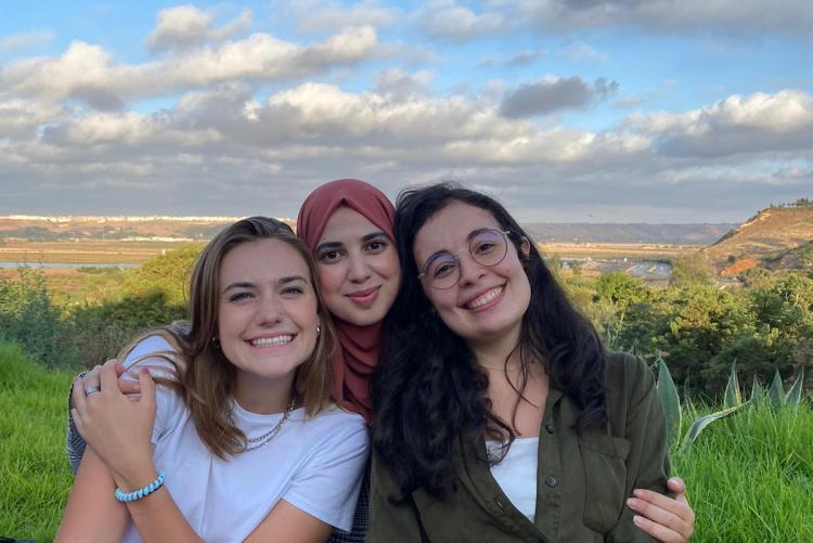 Three women smile and pose for a picture while sitting on the grass during a picnic. The sky is blue and the grass very green.