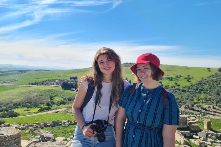 Two young women pose in front of a green landscape scattered with ruins. The sky is bright blue with wispy clouds.