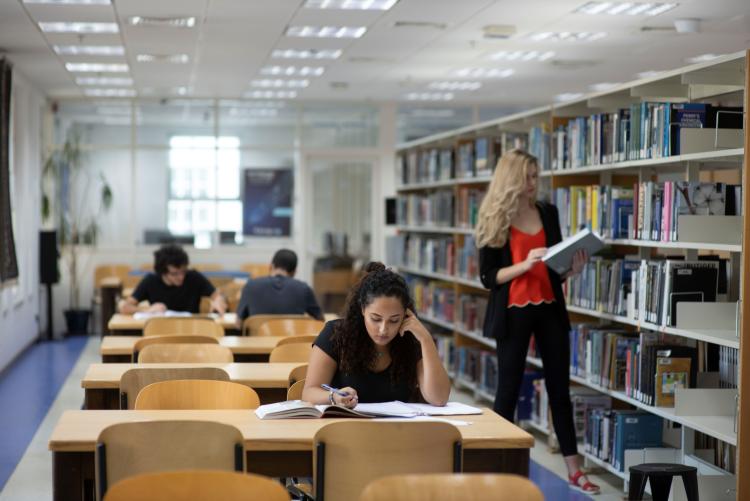 Inside the university library one woman reads a book from off of the shelf while several students sit and study at different tables.