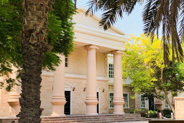 A building on campus with pillars sits in the sun with palm trees in front and around.