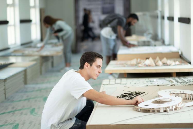 A student crouches on the ground to work on an architectural model placed on a low table. There are several other similar tables in the background.