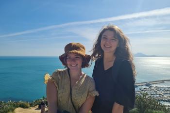 Two women smile from a vantage point that overlooks the blue sea under a sunny blue sky.