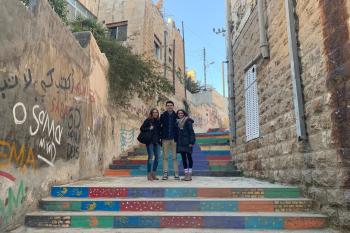 Three students pose on a colorful staircase between buildings in Amman. They are in the middle of the photo on a landing. The wall on the left side has graffiti on it.