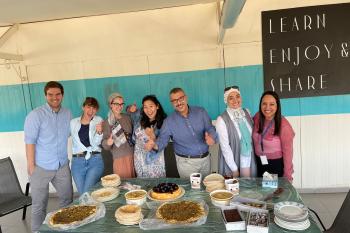 Students stand smiling with their Arabic teachers behind a table laden with many snacks and treats.