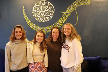 Four women stand together and smile at the camera in front of a dark colored wall with a calligraphy mural on it. 