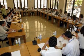 A large group of students sit at multiple long tables facing each other with paper and microphones